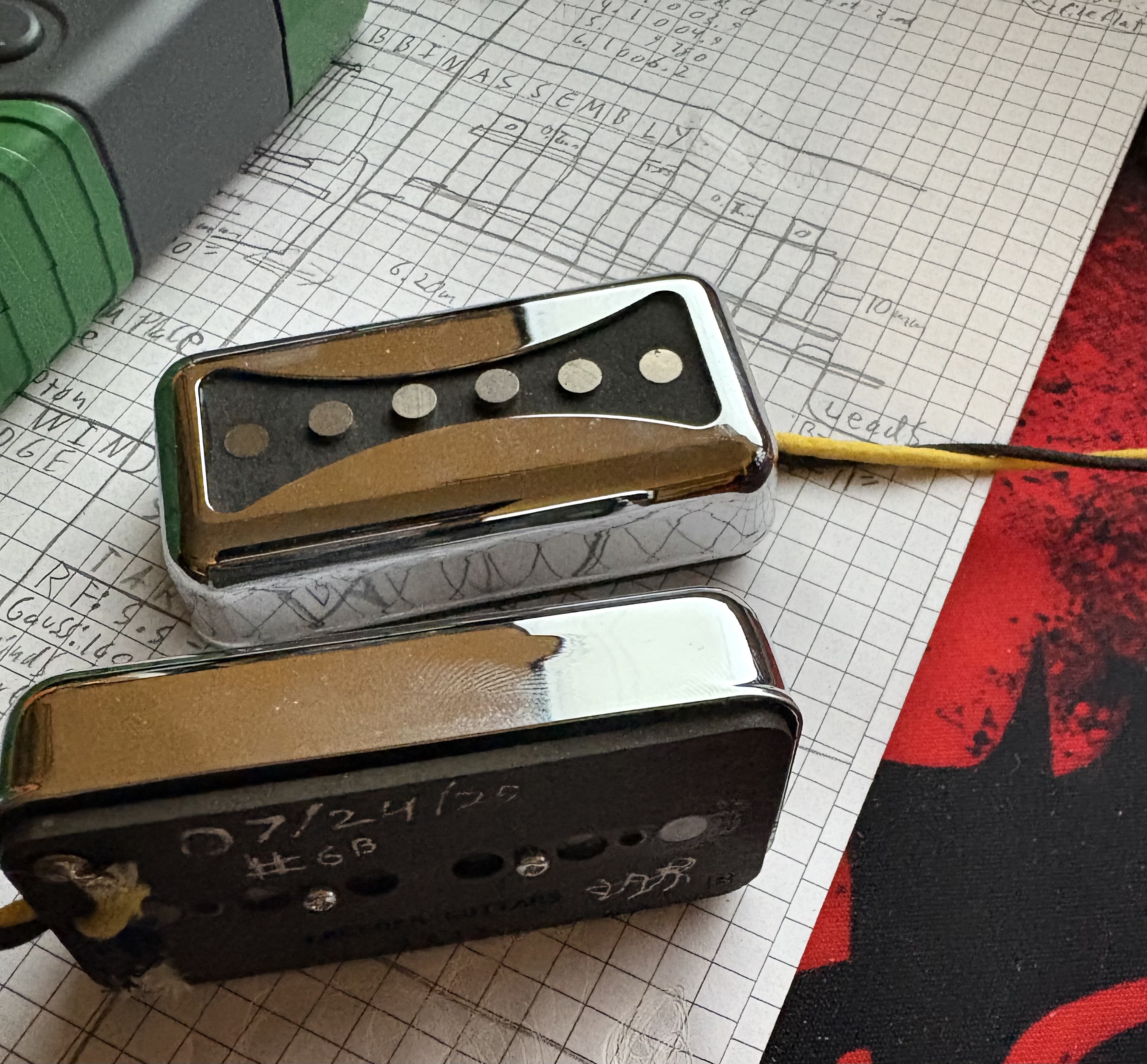 Two finished Arcform Bowtie pickups laid on hand-drawn graph-paper schematic with Zia sun symbol underneath